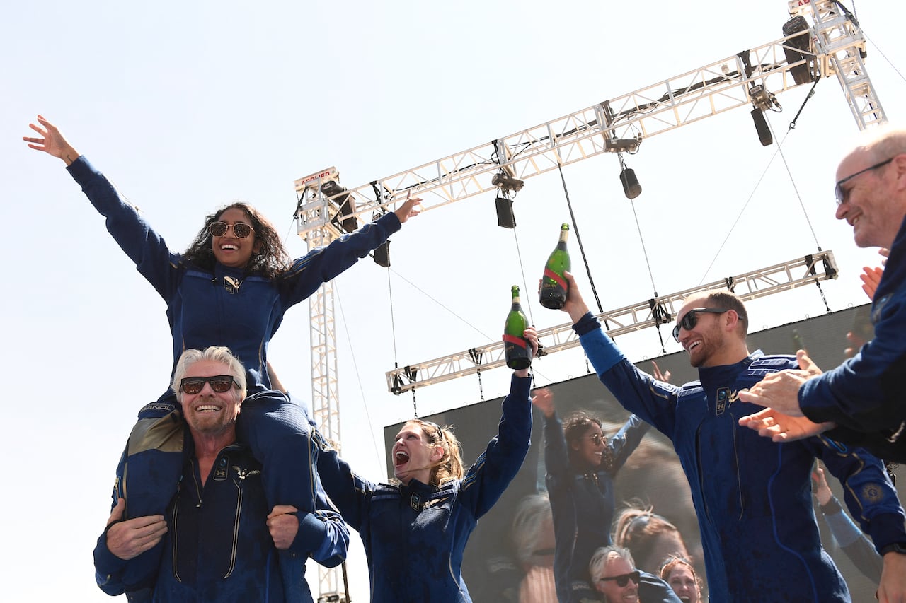 Several people wearing blue jumpsuits celebrate with champagne.