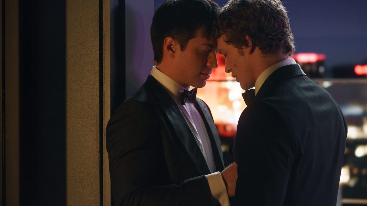 Two men in suits stand with their foreheads touching in front of a window.